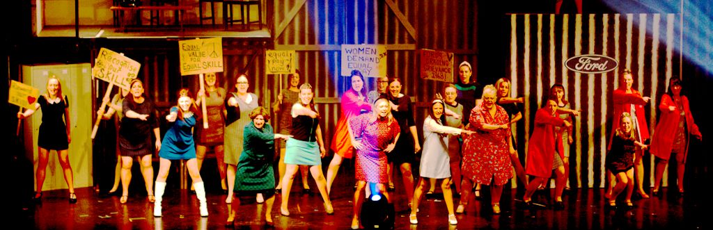 A group of women on stage are holding signs and gesturing, appearing to be part of a protest or reenactment. A Ford logo is visible in the background.