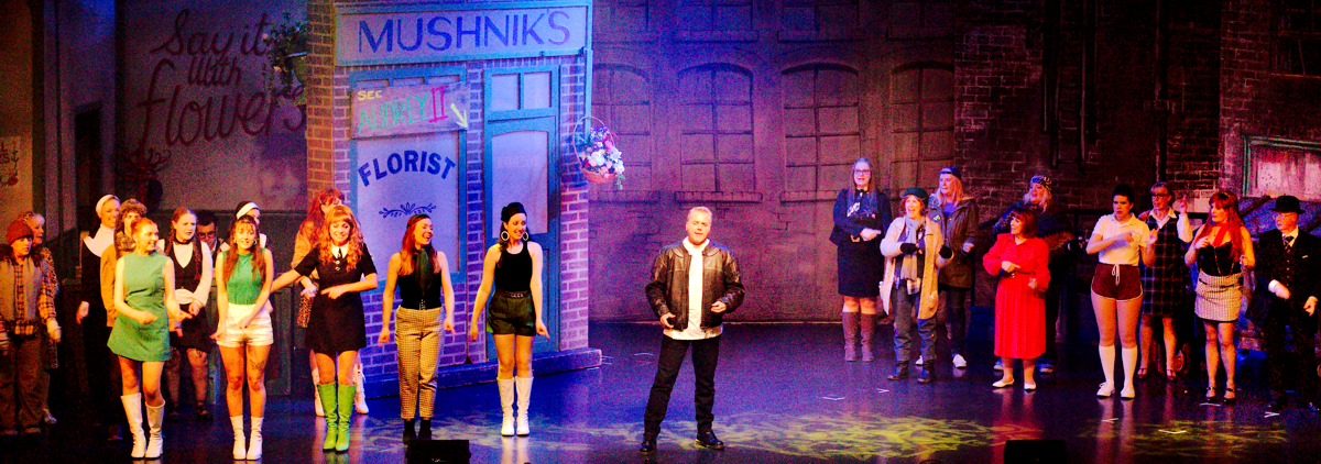 A group of actors on stage in front of a florist shop. A dentist in a black leather jacket stands centre stage. Other actors in colourful costumes surround him.